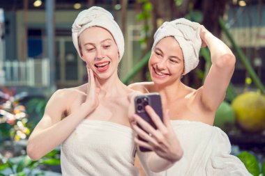 Two Caucasian female friends in towel and head wrap resting relaxing massage in spa, happy friends together taking smartphone selfie in hotel spa salon.wellness, beauty ,spa and relaxation concept