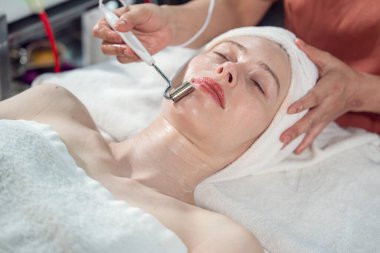 Beautiful young caucasian woman lying on bed having facial massage therapist applying facial cream, moisturizing facial spa in spa salon.