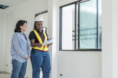 Beyaz kadın müşteri, inşaat miğferi takmış Afrikalı mühendis ile birlikte evi teftiş ediyor. Elinde yeni bir evin dekorasyonu ve dekorasyonu için planlar var. İnşaat ve Emlak kavramı.