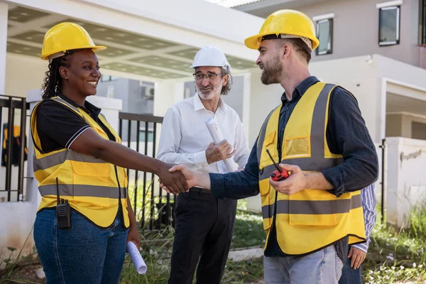 African engineer in construction helmet holding hand and introducing himself, Caucasian male foreman watching construction work together with male executive standing and talking in a housing site