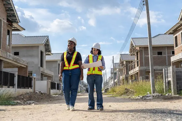 African engineer in construction helmet holding blueprints, renovating a new house, talking with a Caucasian female project looking at construction together in housing site.