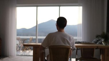 a man is resting in a country house sitting on an armchair and drinking tea, looking at the mountains outside the window