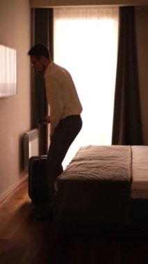  Klasik takım elbiseli genç bir adam elinde bavulla yatak odasına girer ve yatağa oturur.