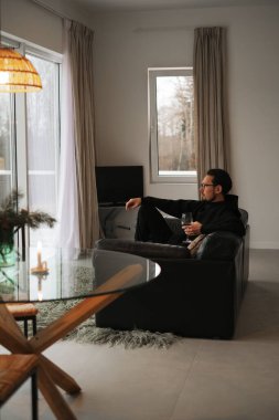 Kanepede oturup panoramik bir evin penceresinden dağlara bakan bir adamın portresi.