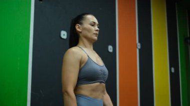 Gri eşofmanlı güzel bayan sporcu spor salonunda kollarını dambıllarla sallıyor.