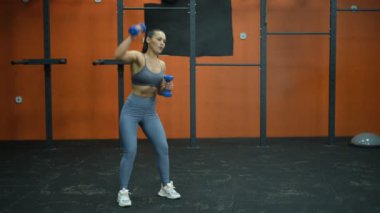 Güzel kaslı sporcu kadın kollarını salonda dambıllarla sallıyor.