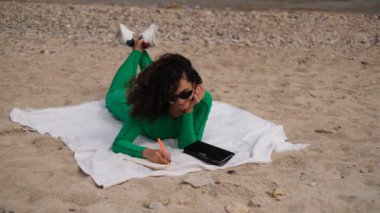 Genç bir kadın plajda uzanıyor ve not defterine notlar yazıyor.