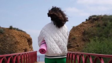 Genç, narin sporcu kadın elinde yoga minderiyle denize doğru yürüyor.