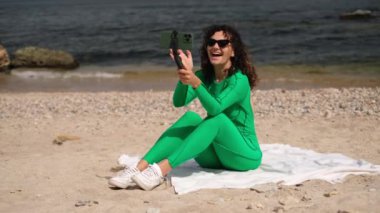 Genç ve güzel bir kadının kumsalda otururken telefonda görüntülü konuşması.