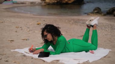 Genç ve güzel bir kadın sahilde yatarken not defterine notlar yazıyor.