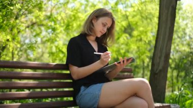 Genç bir kız öğrenci, güneşli bir yaz gününde bir bankta not defterine notlar yazar.