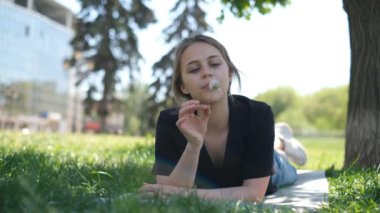 Genç ve güzel bir kız öğrenci çimenlikteki parkta elinde bir karahindiba ile güneşli sıcak bir yaz gününün tadını çıkarıyor.