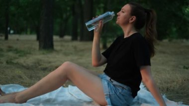Parkta güneşli bir günde piknikte su içen genç bir kadın.