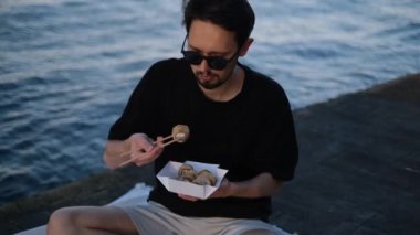 Genç, yakışıklı esmer adam, deniz kıyısındaki bir piknikte Çin yemek çubuklarıyla rulo yiyor.