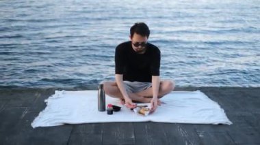 Genç, yakışıklı esmer adam, deniz kıyısındaki bir piknikte Çin yemek çubuklarıyla rulo yiyor.