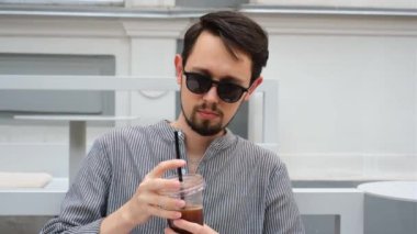 Güneş gözlüklü genç bir adam bir yaz günü terasta plastik bir bardaktan kahve içer.