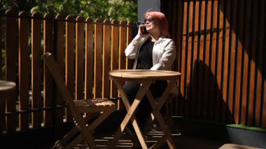 Pembe saçlı şişman genç bir kadın güneşli bir yaz gününde terasta otururken telefonla konuşuyor..