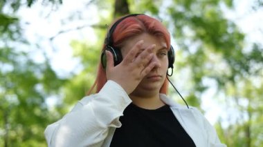 Pembe saçlı şişman genç, güneşli bir günde bir bankta oturmuş kulaklıkla müzik dinliyor.