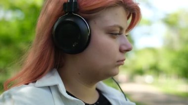 Pembe saçlı şişman genç, güneşli bir günde bir bankta oturmuş kulaklıkla müzik dinliyor.