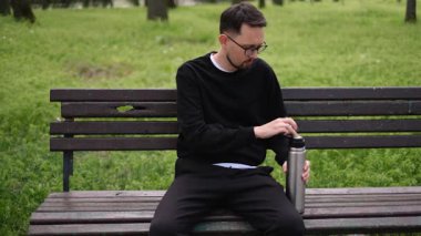 Genç adam parkta bankta otururken termostan fincana çay dolduruyor.