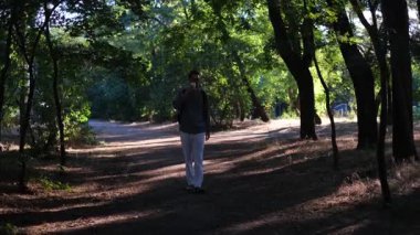 Genç yakışıklı bir adam güneşli bir yaz sabahı parkta bir fincan kahveyle yürüyor.