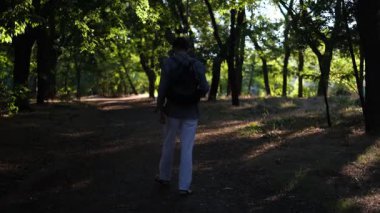 Genç yakışıklı bir adam güneşli bir yaz sabahı parkta bir fincan kahveyle yürüyor.