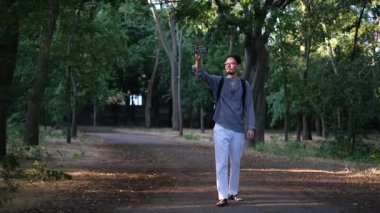 Genç bir blogcu güneşli bir yaz sabahı parkta yürüyor ve video kaydediyor.