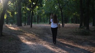 Genç bir kadın parkta yürüyor ve yaz günü şafak vakti doğayı telefonuyla filme alıyor.
