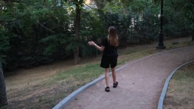 Genç bir kadın sıcak bir yaz akşamı telefonda konuşurken parkta yürüyor.