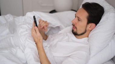 Sakallı genç esmer uyandı ve yatakta yatıp telefonundan haberleri izledi.
