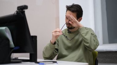 Genç bir adam bilgisayar masasında oturuyor, gözlüklerini çıkarıyor ve gözlerini yorgunluktan ovalıyor.