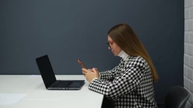 Ofiste otururken telefonla mesajlaşan gözlüklü genç kadın.