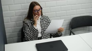 Ofisteki masasında telefonla konuşurken rapor okuyan genç bir kadın.