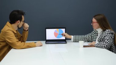 Genç iş ortakları ofiste karşılıklı oturup dizüstü bilgisayara bakarak bir anlaşma yapıyorlar.