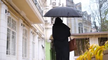 Sonbahar yağmurlu günlerinde şehir merkezinde şemsiyeyle yürüyen siyah paltolu genç bir kadın mimariye bakıyor.