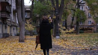 Siyah ceketli genç bir kadın telefonda video çekip yağmurlu sonbahar günlerinde mimariye bakıyor.