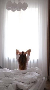 Genç bir kadın, güneşli bir sabah uyandıktan sonra uzanarak yatağın kenarına oturur.