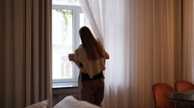 Genç bir kadın sabahın erken saatlerinde evde çay yapıp pencereden dışarı bakıyor.