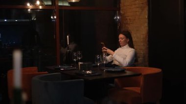 Uzun saçlı genç bir kadın restoran masasında yalnız başına yemek için kendisine eşlik edecek birini bekliyor.