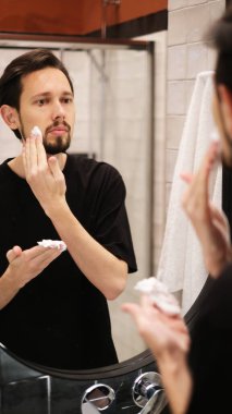 Genç adam aynaya bakarken yanaklarına tıraş köpüğü sürüyor.