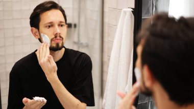Genç adam aynaya bakarken yanaklarına tıraş köpüğü sürüyor.