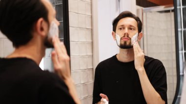 Genç adam aynaya bakarken yanaklarına tıraş köpüğü sürüyor.