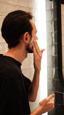 Genç adam aynaya bakarken yanaklarına tıraş köpüğü sürüyor.