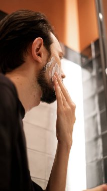 Genç adam aynaya bakarken yanaklarına tıraş köpüğü sürüyor.