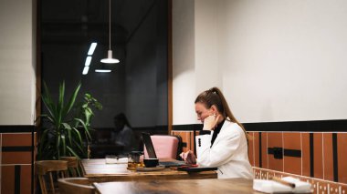 Beyaz ceketli güzel bir kadın kafede dizüstü bilgisayarla çalışıyor.