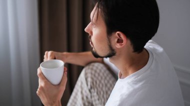 Genç esmer adam yatağın kenarında oturuyor, huzurlu bir sabahta pencereden dışarı bakarken çayını yudumluyor.