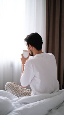 Genç esmer adam yatağın kenarında oturuyor, huzurlu bir sabahta pencereden dışarı bakarken çayını yudumluyor.