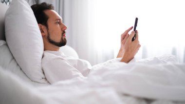 Genç esmer adam sabahın erken saatlerinde parlak, rahat bir yatak odasında telefonuna bakıyor.