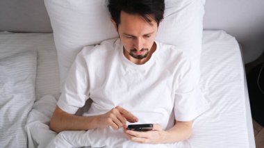 Genç esmer adam sabahın erken saatlerinde parlak, rahat bir yatak odasında telefonuna bakıyor.