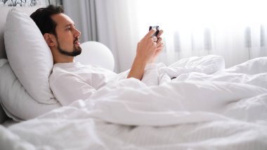 Genç esmer adam sabahın erken saatlerinde parlak, rahat bir yatak odasında telefonuna bakıyor.